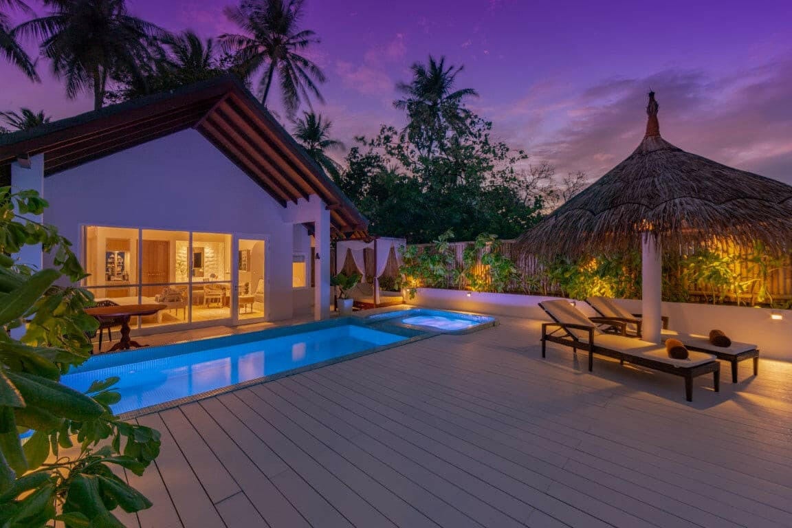 A villa at Malahini Kuda Bandos lit at dusk, its pool and thatched sala on the deck