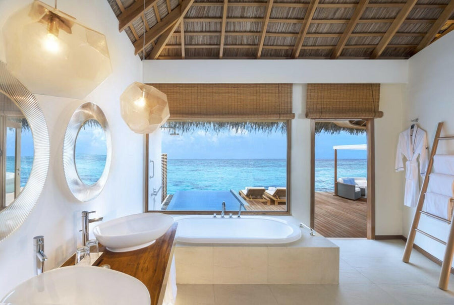A villa bathroom at W Maldives, porthole windows and an oval tub facing the open lagoon.