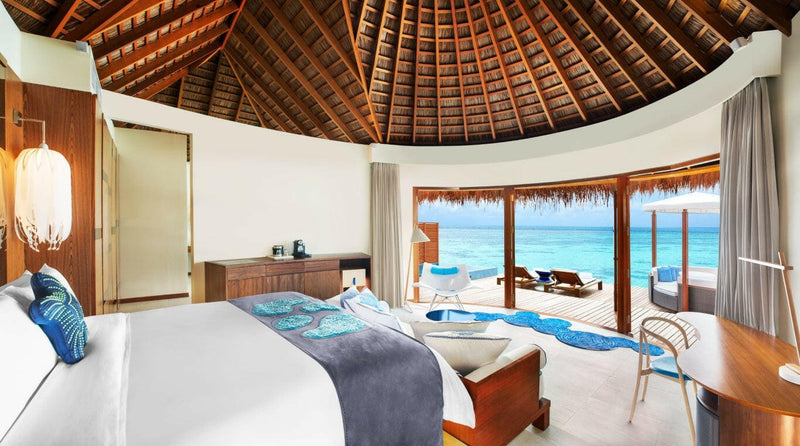 A bedroom at W Maldives under a round timber roof, grey runner and turquoise cushions on the bed.