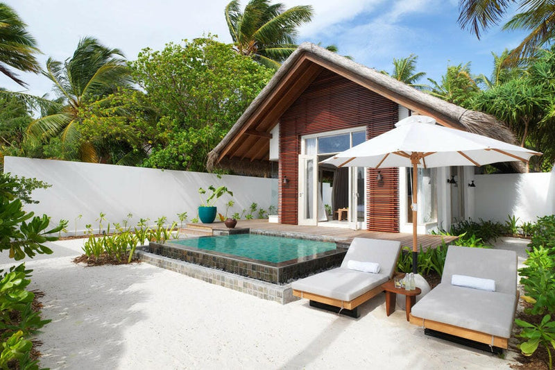 A beach villa at Sirru Fen Fushi, grey loungers and a timber parasol beside a dark edged pool.