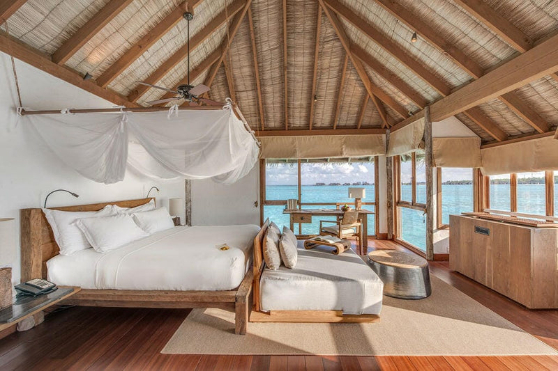 A residence bedroom at Gili Lankanfushi, curtained four-poster bed and timber windows onto the water.