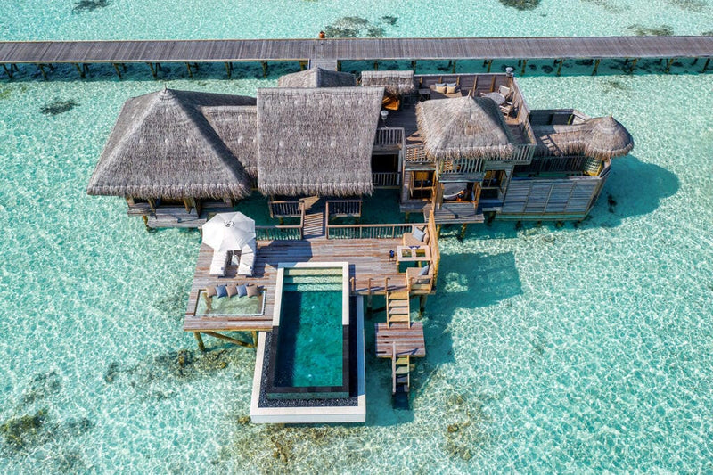 Gili Lankanfushi from the air, thatched roofs around a rectangular pool and a timber jetty.
