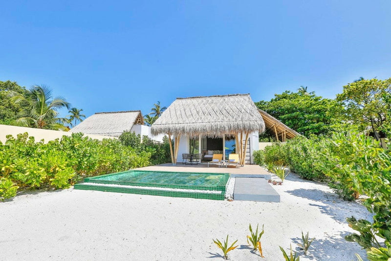 A beach villa at Emerald Maldives, green tiled pool on white sand in front of a thatched pavilion.