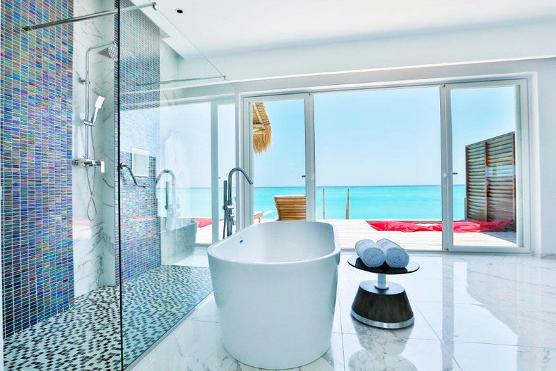 A villa bathroom at Emerald Maldives, oval tub beside a blue mosaic shower and doors to the sea.