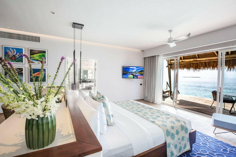 A villa bedroom at Emerald Maldives, white lilies on a ledge and doors open to a deck and the sea.