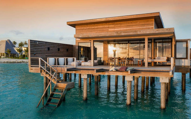 An overwater villa at Kuda Villingili on timber stilts, glass walls glowing at golden hour.