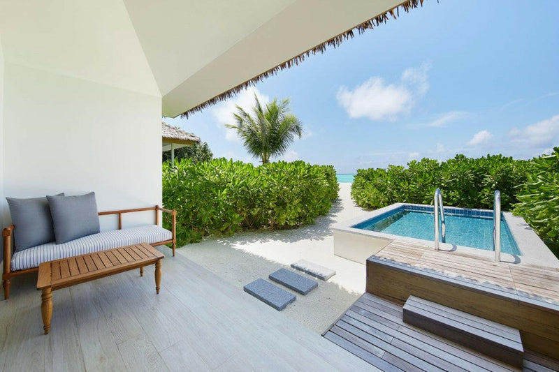 A villa terrace at Le Meridien Maldives, timber sofa under thatch beside a hedged rectangular pool.