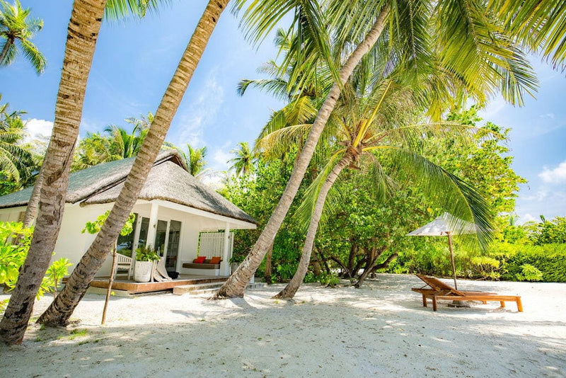 A white villa at LUX* South Ari Atoll among leaning coconut palms, lounger and parasol on the sand.