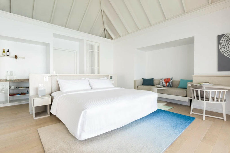 A villa bedroom at LUX* South Ari Atoll, white walls and a blue rug beside a pale seating corner.