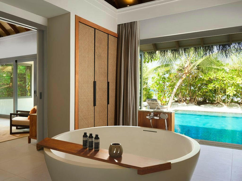 A villa bathroom at Vakkaru Maldives, oval freestanding tub with a timber tray, pool and greenery outside.