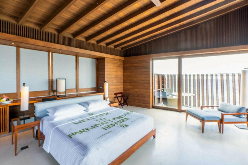 An overwater villa bedroom at Park Hyatt Maldives Hadahaa, a white bed in a timber lined room opening to the deck