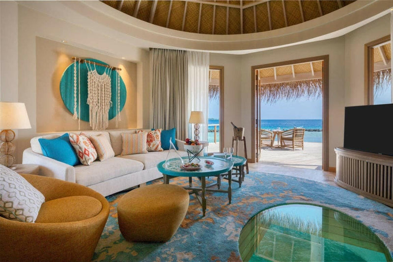 The living room of a house at The Nautilus Maldives, cream sofas and tan armchairs with the doors open to the sea