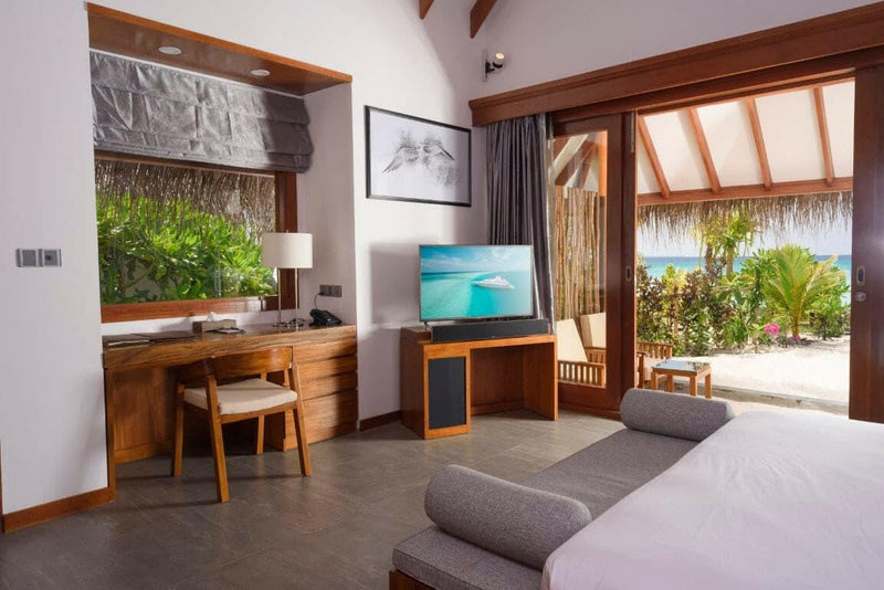A beach villa bedroom at Dhigufaru Island Resort, timber desk and doors open to a palm garden.