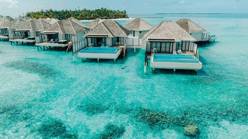 A row of water villas at Nova Maldives with dark reef patches showing through the shallow lagoon.