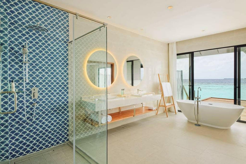 A villa bathroom at Nova Maldives, blue mosaic shower behind glass and a freestanding tub by the window.
