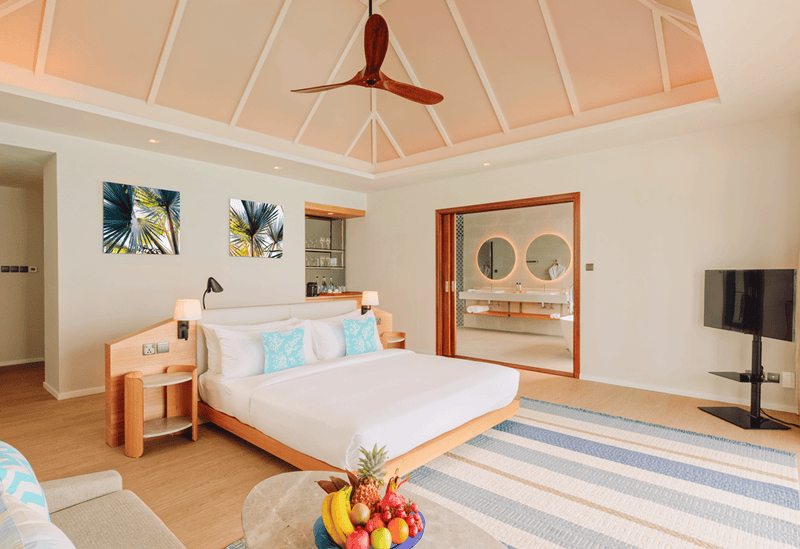 A bedroom at Nova Maldives, white bed with turquoise cushions and a doorway to twin round mirrors.