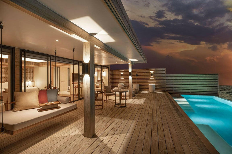 The deck of a water villa at Kuda Villingili at dusk, a daybed and a lit pool under a dark sky.
