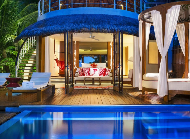 A lit villa at W Maldives after dark, blue canopied daybed on the deck above a glowing pool.
