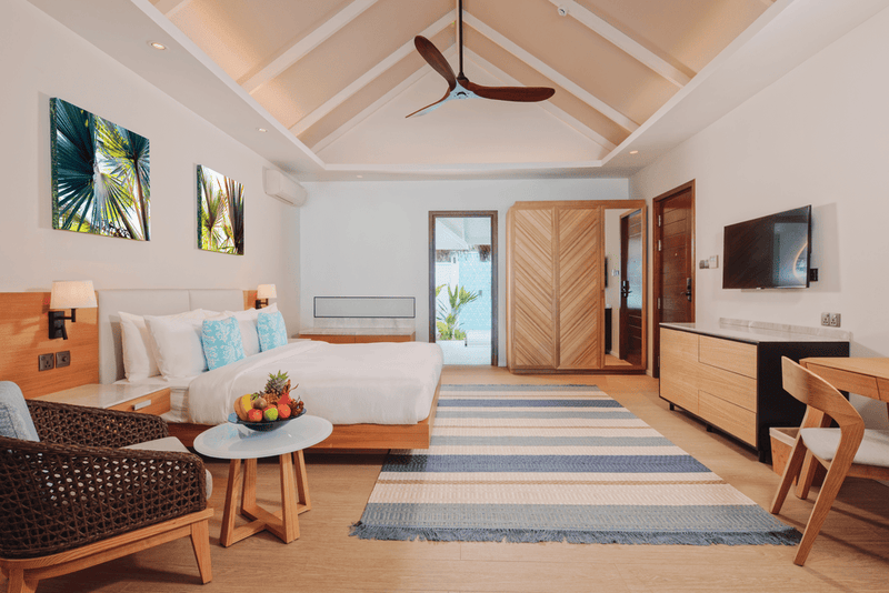 A bedroom at Nova Maldives seen from the door, rattan armchair, fruit bowl and framed palm prints.